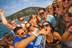 Group of friends partying on a private Budapest booze cruise, energetic onboard atmosphere with Gellért Hill in the background.