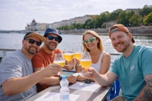 Group of friends toasting with beers on a private Budapest Danube cruise, riverside promenade and city skyline in the background