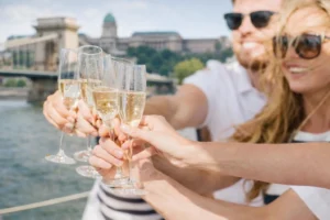 Friends holding prosecco glasses on a boat during a river cruise, with the Chain bridge and the Castle hill behind.