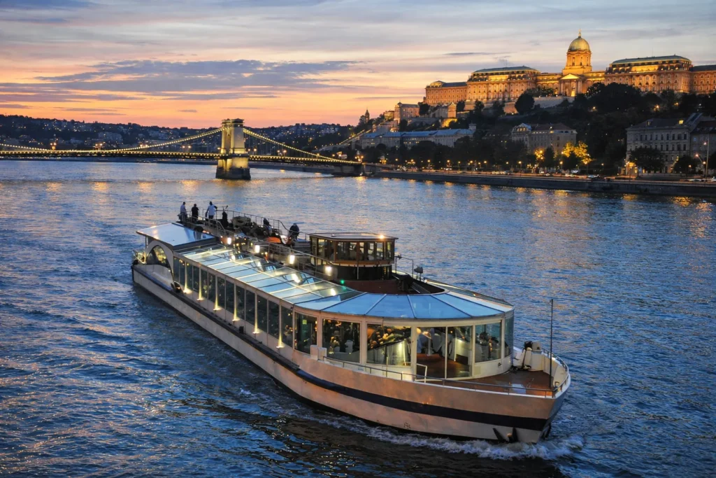 Glass roof river cruise boat on the Danube at dusk in Budapest, with Chain Bridge and Buda Castle glowing in the background.