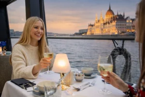 Two friends enjoying white wine on a Budapest dinner cruise at sunset, with the Hungarian Parliament visible across the Danube.