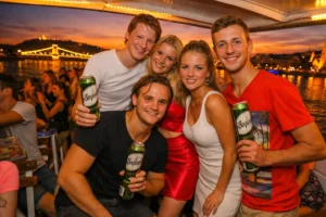 Group of friends enjoying a sunset party cruise on the Danube in Budapest, holding beers on board with warm evening lights and river views.