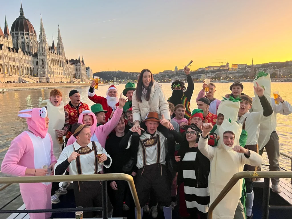 Costumed group celebrating on a boat in Budapest at sunset, with the Hungarian Parliament and the Danube in the background.