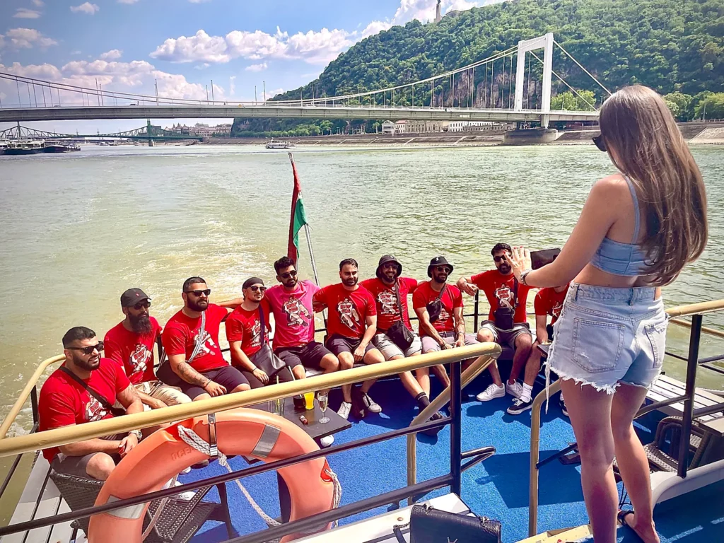 Group photo on a boat in Budapest on the Danube, friends in matching shirts with Liberty Bridge and the riverside hills in the background.