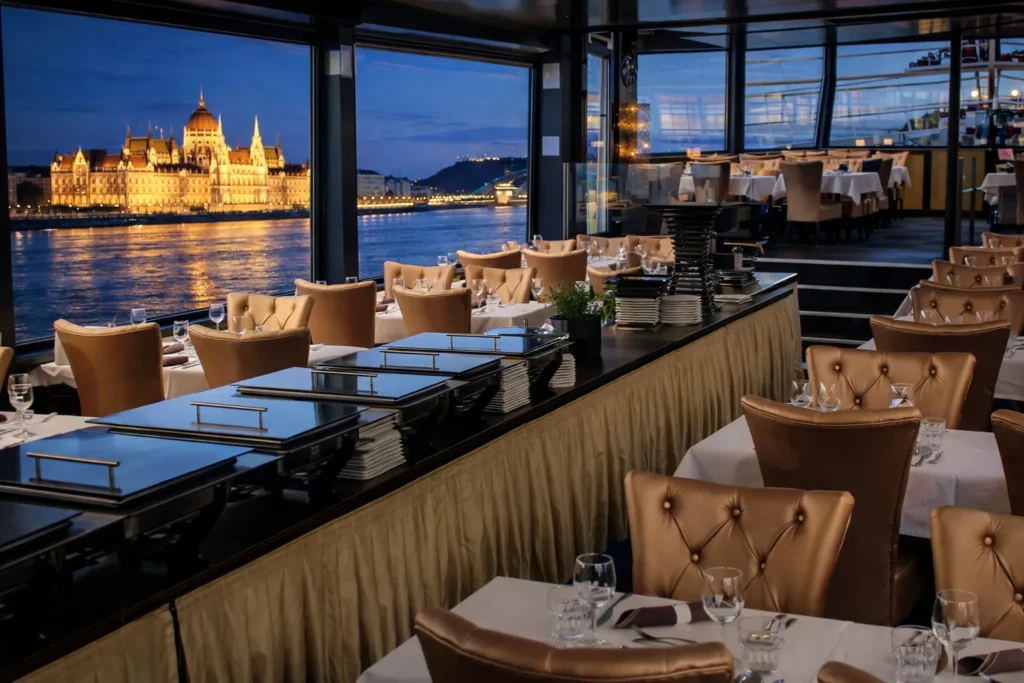 Warm indoor dining lounge on a private boat with large windows looking out to the Danube and Budapest Parliament at dusk, indoor setup option for a private cruise.